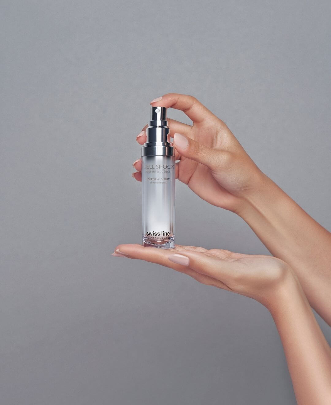 Sérum Essentiel Cell Shock Swissline tenu entre deux mains, soin visage anti-âge Age Intelligence sur fond gris doux.